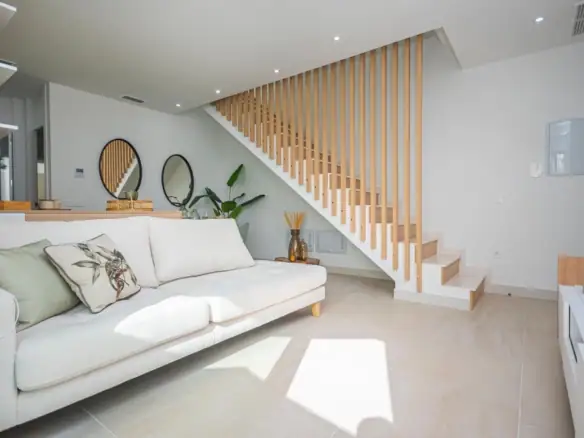 Moderne woonkamer met witte bank, houten trap en groene planten, veel natuurlijk licht en strakke af.